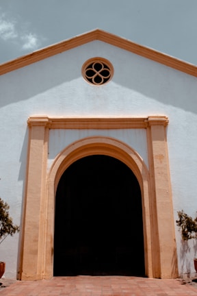 A facade of a historic building with white walls and a large wooden doorway framed by a light brown arch. Above the doorway, there is a small circular window with a decorative design. The building has a simple, symmetrical architecture with soft shadows cast on its surface.