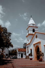 A colonial-style church with a white facade and orange trim stands prominently. The church tower has a clock and a cross at its peak. In the foreground, there are large planters with green shrubs. The plaza is made of reddish-brown tiles, and there's a black car parked nearby. Surrounding buildings are low-rise, with a mix of colonial white walls and red-tiled roofs. A few people are walking in the distance under a partly cloudy sky.