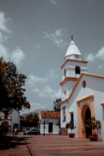 A colonial-style church with a white facade and orange trim stands prominently. The church tower has a clock and a cross at its peak. In the foreground, there are large planters with green shrubs. The plaza is made of reddish-brown tiles, and there's a black car parked nearby. Surrounding buildings are low-rise, with a mix of colonial white walls and red-tiled roofs. A few people are walking in the distance under a partly cloudy sky.