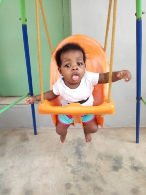 A young child is sitting in an orange swing designed for infants, secured with a safety strap. The swing is attached to a colorful frame with yellow ropes. The child is wearing a white shirt and colorful shorts, looking directly at the camera with an expression of curiosity.