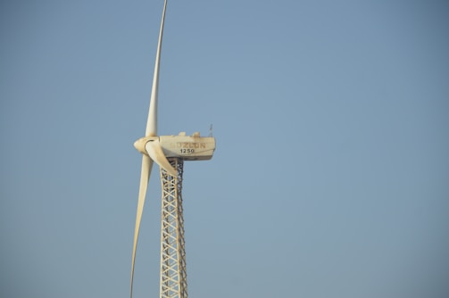 A tall wind turbine with three blades is set against a clear blue sky. The equipment is labeled with 'Solon 1250' on the nacelle, which is mounted on a lattice-style tower.