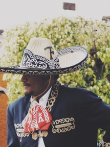 A person wearing traditional Mexican attire including a large, ornate sombrero with intricate patterns and a decorative embroidered charro suit, standing against a background of green foliage.