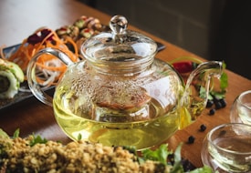 A glass teapot filled with a light green tea is placed on a wooden table. Next to the teapot, there are two small glass cups, partially filled with tea. In the background, various types of sushi and garnishes, including carrot strips and leafy greens, are displayed on a black platter. Black berries are scattered around, enhancing the presentation.