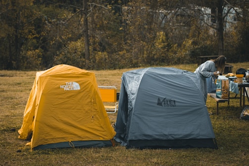 Two camping tents, one yellow and one gray, are pitched on grassy ground. A person stands nearby at a table, likely preparing a meal or organizing items under a canopy of trees with autumn foliage.