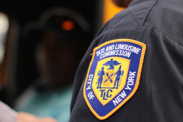 A close-up view of a patch on a uniform, reading 'Taxi and Limousine Commission City of New York' with a yellow and blue emblem showing various figures and symbols.