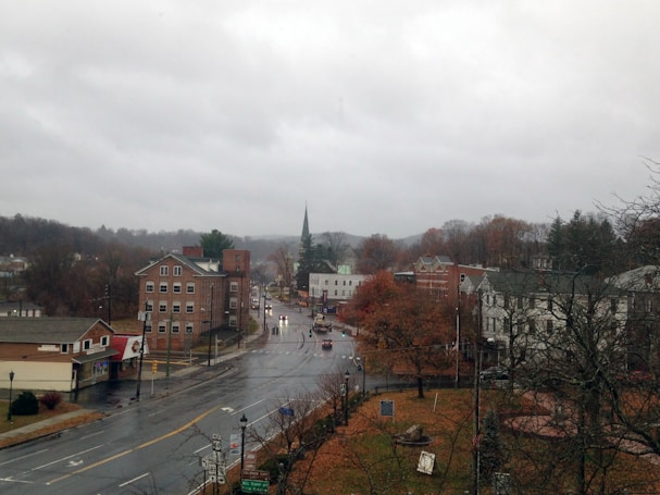 A small town scene featuring several brick buildings, a church with a tall steeple, and autumnal trees lining the street. The sky is overcast, suggesting a rainy or dreary day. The streets appear wet, and there are visible street lamps and road signs. The town is surrounded by a landscape of rolling hills.