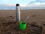 A metal thermos and a green cup are placed on a sandy beach with a cloudy sky and ocean waves in the background. The cup contains what appears to be yerba mate with a metal straw.