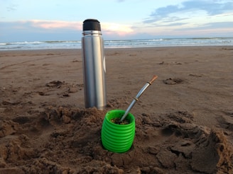 A metal thermos and a green cup are placed on a sandy beach with a cloudy sky and ocean waves in the background. The cup contains what appears to be yerba mate with a metal straw.