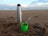 A metal thermos and a green cup are placed on a sandy beach with a cloudy sky and ocean waves in the background. The cup contains what appears to be yerba mate with a metal straw.