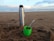 A metal thermos and a green cup are placed on a sandy beach with a cloudy sky and ocean waves in the background. The cup contains what appears to be yerba mate with a metal straw.