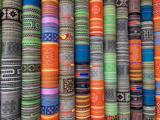 Vibrant rolls of fabric with intricate, multicolored geometric and floral patterns arranged vertically. The patterns vary across different cultures and styles, creating a visually striking display.