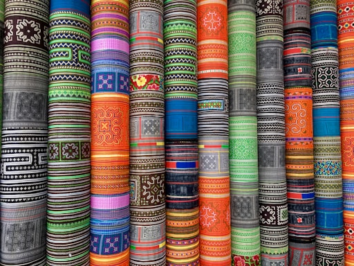 Vibrant rolls of fabric with intricate, multicolored geometric and floral patterns arranged vertically. The patterns vary across different cultures and styles, creating a visually striking display.