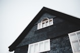 The upper portion of a dark-colored house with a triangular roof is visible. There is a small, arched window with white framing near the peak of the roof and a larger set of rectangular windows below it. The house exterior is covered with dark shingles, and the sky in the background is overcast.