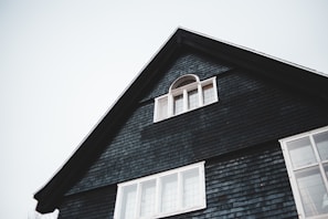 The upper portion of a dark-colored house with a triangular roof is visible. There is a small, arched window with white framing near the peak of the roof and a larger set of rectangular windows below it. The house exterior is covered with dark shingles, and the sky in the background is overcast.