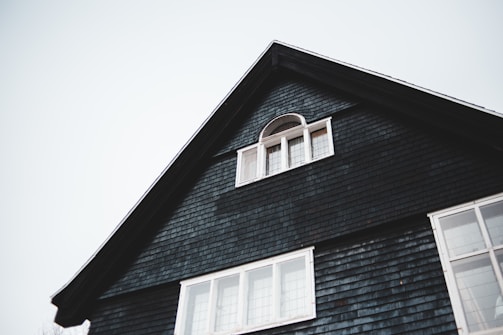 The upper portion of a dark-colored house with a triangular roof is visible. There is a small, arched window with white framing near the peak of the roof and a larger set of rectangular windows below it. The house exterior is covered with dark shingles, and the sky in the background is overcast.
