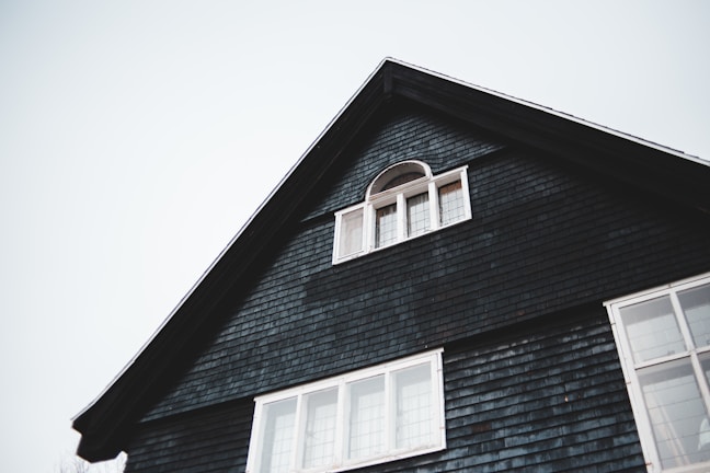 The upper portion of a dark-colored house with a triangular roof is visible. There is a small, arched window with white framing near the peak of the roof and a larger set of rectangular windows below it. The house exterior is covered with dark shingles, and the sky in the background is overcast.