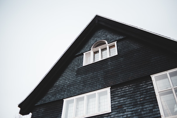 The upper portion of a dark-colored house with a triangular roof is visible. There is a small, arched window with white framing near the peak of the roof and a larger set of rectangular windows below it. The house exterior is covered with dark shingles, and the sky in the background is overcast.