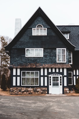 A classic two-story house with a steeply pitched roof and dark, shingle-covered walls. The facade features a combination of stone and timber accents, with a prominent white wooden door and several rectangular windows. Surrounding the house are some trees and a paved path in the foreground.