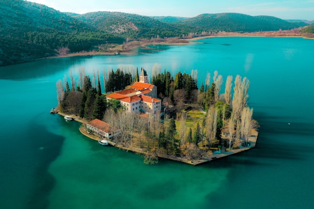 A scenic island is surrounded by a serene lake with clear turquoise water. The island hosts a historical building with red roofs, nestled among tall trees, including cypress and poplar. The landscape in the background consists of lush green hills under a clear blue sky.