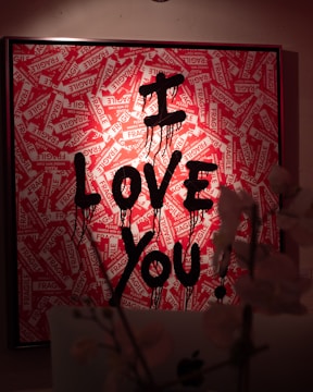 A framed artwork features red and white 'Fragile' stickers arranged in various directions as a background. In front of these stickers, the words 'I LOVE YOU' are written in bold, dripping black paint. The lighting casts a warm glow, and there are blurred plants or flowers in the foreground.