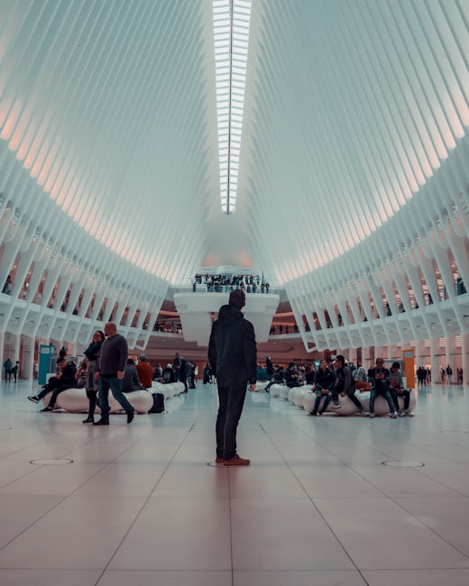 A modern architectural space with a high, vaulted ceiling composed of repeating white beams converging at a central skylight. People are gathered in various groups throughout the expansive, open area, some sitting on white benches while others stand or walk. The interior lighting creates a soft, ambient atmosphere.