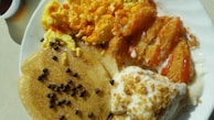A plate filled with various breakfast foods including scrambled eggs, breaded chicken tenders with sauce, a pancake topped with chocolate chips, and a portion of yogurt with granola. The meal is arranged on a round white plate under natural light.
