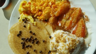 A plate filled with various breakfast foods including scrambled eggs, breaded chicken tenders with sauce, a pancake topped with chocolate chips, and a portion of yogurt with granola. The meal is arranged on a round white plate under natural light.