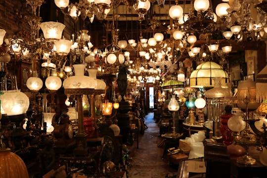 A densely packed shop displays an array of antique lamps and chandeliers with ornate designs and various styles. The lighting creates a warm, inviting glow that highlights the intricate details of the lamps. The narrow aisle is filled with both table lamps and hanging chandeliers, contributing to a sense of timelessness and nostalgia.