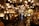 A densely packed shop displays an array of antique lamps and chandeliers with ornate designs and various styles. The lighting creates a warm, inviting glow that highlights the intricate details of the lamps. The narrow aisle is filled with both table lamps and hanging chandeliers, contributing to a sense of timelessness and nostalgia.