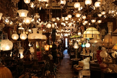 A densely packed shop displays an array of antique lamps and chandeliers with ornate designs and various styles. The lighting creates a warm, inviting glow that highlights the intricate details of the lamps. The narrow aisle is filled with both table lamps and hanging chandeliers, contributing to a sense of timelessness and nostalgia.