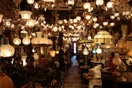 A densely packed shop displays an array of antique lamps and chandeliers with ornate designs and various styles. The lighting creates a warm, inviting glow that highlights the intricate details of the lamps. The narrow aisle is filled with both table lamps and hanging chandeliers, contributing to a sense of timelessness and nostalgia.