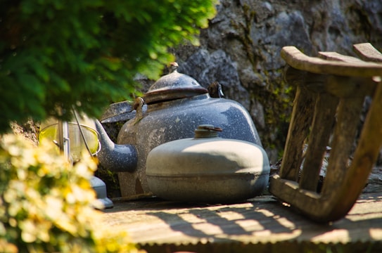 An old metal kettle and other vintage cooking pots are placed on a wooden surface outdoors, partially shaded by lush green foliage. The background features a textured stone wall, enhancing the rustic and nostalgic atmosphere.