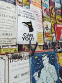 Various vintage and retro-style metal signs are displayed closely together. The signs feature different graphics including a dog with the message 'I CAN MAKE IT TO THE FENCE IN 2.8 SECONDS. CAN YOU?', comic book covers, ads with humorous and motivational texts, and popular culture references. The signs have a worn and rusty appearance, suggesting age.