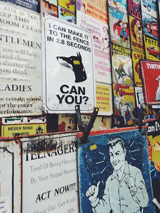 Various vintage and retro-style metal signs are displayed closely together. The signs feature different graphics including a dog with the message 'I CAN MAKE IT TO THE FENCE IN 2.8 SECONDS. CAN YOU?', comic book covers, ads with humorous and motivational texts, and popular culture references. The signs have a worn and rusty appearance, suggesting age.