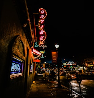 A vibrant street scene at night features colorful neon signs advertising a lounge. The sign reads 'Satire Lounge' with a martini glass icon, and the storefronts are illuminated, reflecting a lively atmosphere. Cars line the well-lit road, and a few pedestrians are visible under the streetlights.