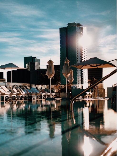A modern urban setting with a rooftop pool surrounded by lounge chairs and umbrellas. High-rise buildings can be seen in the background, and the sun is casting reflections on the water, creating a serene and luxurious atmosphere.