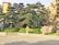 A meticulously landscaped garden featuring carefully trimmed trees with lush, rounded canopies. Large ornamental rocks are strategically placed throughout the garden, which is enclosed by a low stone fence. The setting is sunlit, casting soft shadows on the neatly paved ground.