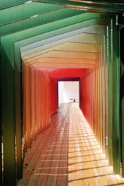 A walkway framed by a series of colored panels transitioning from green to red, creating a tunnel effect with alternating light and shadow patterns on a wooden floor. The end of the tunnel reveals a brightly lit, white exit with a small fire extinguisher mounted on the wall.