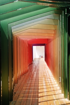 A walkway framed by a series of colored panels transitioning from green to red, creating a tunnel effect with alternating light and shadow patterns on a wooden floor. The end of the tunnel reveals a brightly lit, white exit with a small fire extinguisher mounted on the wall.
