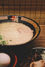 A bowl of creamy ramen sits on a wooden table, with a dark bowl adorned with red and gold patterns on the rim. The ramen features a light-colored broth and thin noodles, topped with a slice of meat and garnished with green onions. In the foreground, there is an egg in a separate bowl and other ingredients.