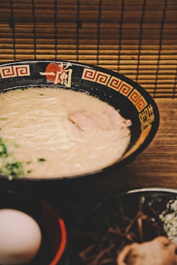 A bowl of creamy ramen sits on a wooden table, with a dark bowl adorned with red and gold patterns on the rim. The ramen features a light-colored broth and thin noodles, topped with a slice of meat and garnished with green onions. In the foreground, there is an egg in a separate bowl and other ingredients.