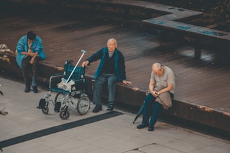 Three elderly individuals are seated on a wooden platform. One person sits next to an unoccupied wheelchair with a crutch resting against it, while another person appears engrossed in their phone. The setting is outdoors, with a tiled floor in the foreground and benches behind them.