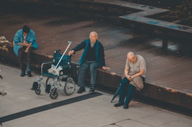 Three elderly individuals are seated on a wooden platform. One person sits next to an unoccupied wheelchair with a crutch resting against it, while another person appears engrossed in their phone. The setting is outdoors, with a tiled floor in the foreground and benches behind them.