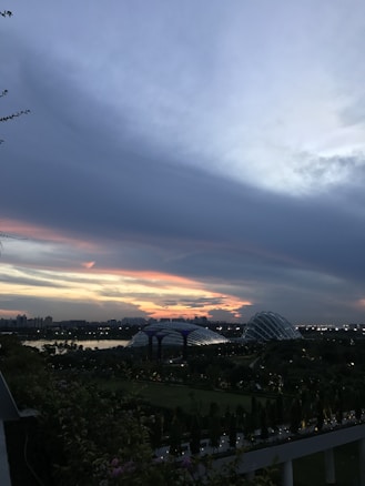 A twilight scene featuring a cityscape with silhouetted buildings against a vibrant sunset sky filled with dynamic clouds. The foreground includes lush greenery and a distinctive futuristic structure with a glass dome. City lights begin to illuminate, creating an enchanting evening atmosphere.