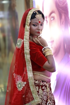 A woman is dressed in traditional Indian attire featuring a red and gold saree with intricate embroidery. She wears matching gold jewelry, including bangles, a necklace, and a nose ring. Her makeup is detailed with a noticeable red bindi on her forehead. The background is softly blurred with a poster or picture.