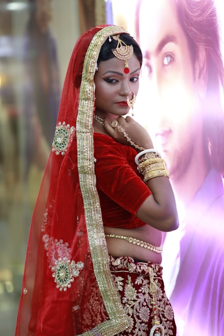 A woman is dressed in traditional Indian attire featuring a red and gold saree with intricate embroidery. She wears matching gold jewelry, including bangles, a necklace, and a nose ring. Her makeup is detailed with a noticeable red bindi on her forehead. The background is softly blurred with a poster or picture.