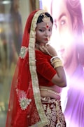 A woman is dressed in traditional Indian attire featuring a red and gold saree with intricate embroidery. She wears matching gold jewelry, including bangles, a necklace, and a nose ring. Her makeup is detailed with a noticeable red bindi on her forehead. The background is softly blurred with a poster or picture.