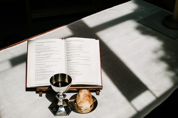 An open book with visible text, a silver chalice containing red liquid, and a piece of bread are arranged on a white tablecloth. Soft shadow lines cross the surface, creating a serene atmosphere.