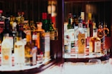A well-lit bar shelf displaying an assorted collection of liquor bottles, including whiskey, vodka, and other spirits, organized neatly with some glassware placed in front. The atmosphere suggests a cozy, inviting setting.