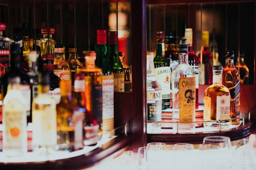 A well-lit bar shelf displaying an assorted collection of liquor bottles, including whiskey, vodka, and other spirits, organized neatly with some glassware placed in front. The atmosphere suggests a cozy, inviting setting.